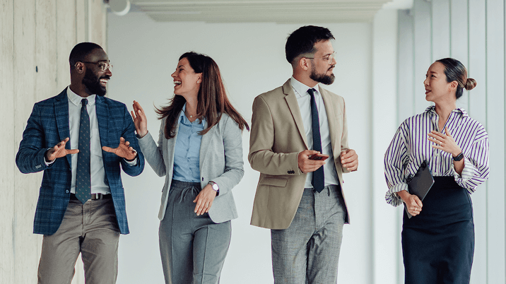 多様な国籍の男女4人のビジネスパーソンが、オフィスの廊下を歩きながら楽しそうに会話している